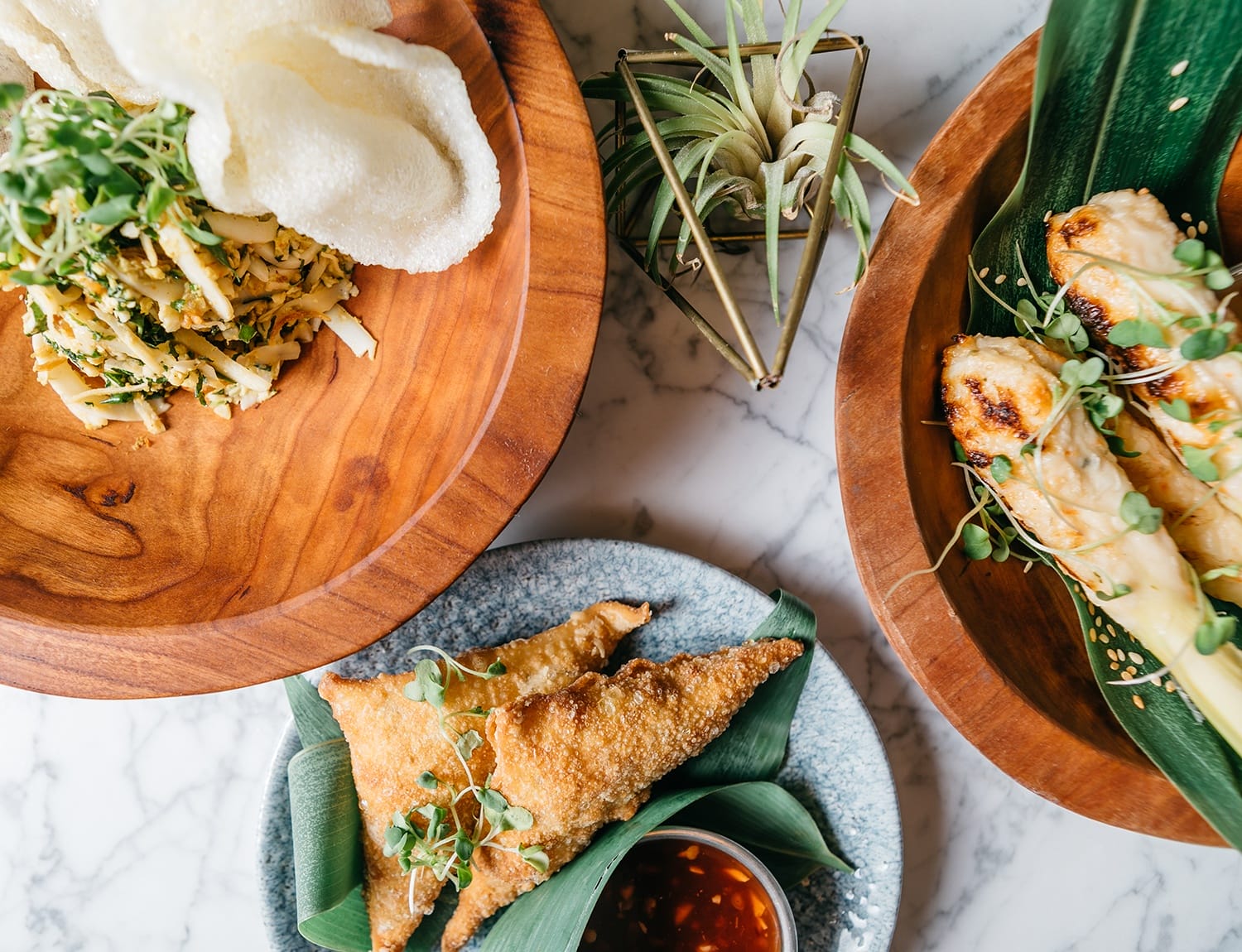 Top view of diverse Asian dishes on wooden plates, including crispy snacks, grilled skewers, and a small plant on marble surface.