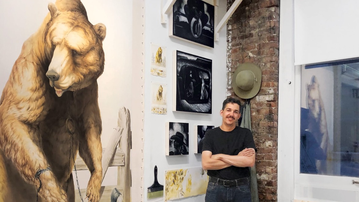Artist standing in a studio with arms crossed, surrounded by various paintings and a large bear artwork on the wall.