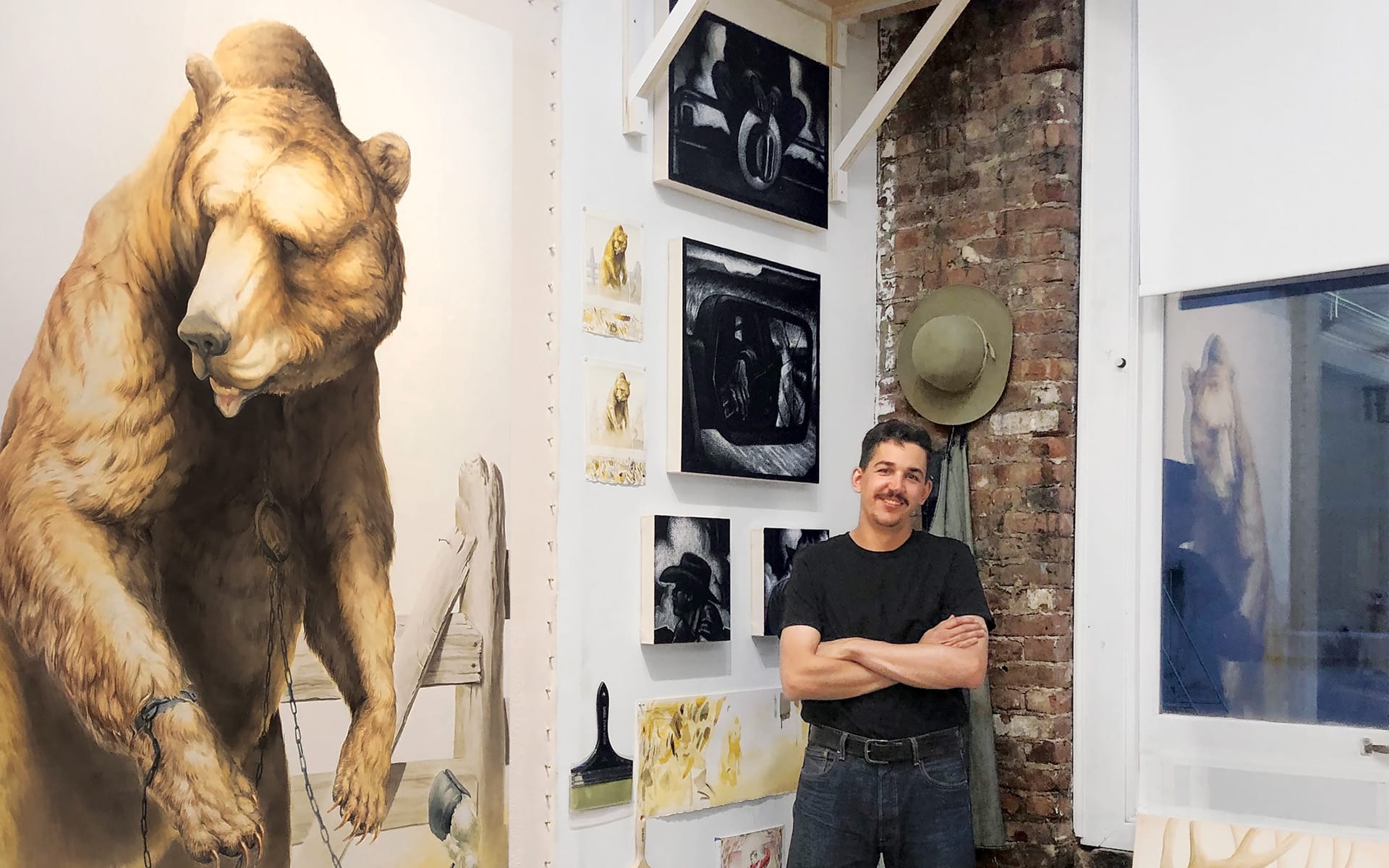 Artist standing in a studio with arms crossed, surrounded by various paintings and a large bear artwork on the wall.