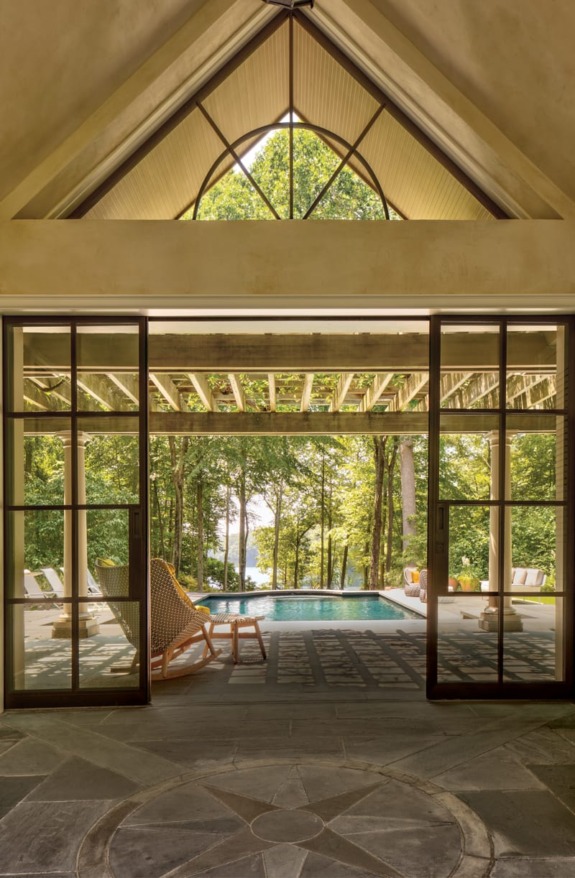 View through large glass doors of a serene patio with a pool, surrounded by lush trees and cushioned seating.