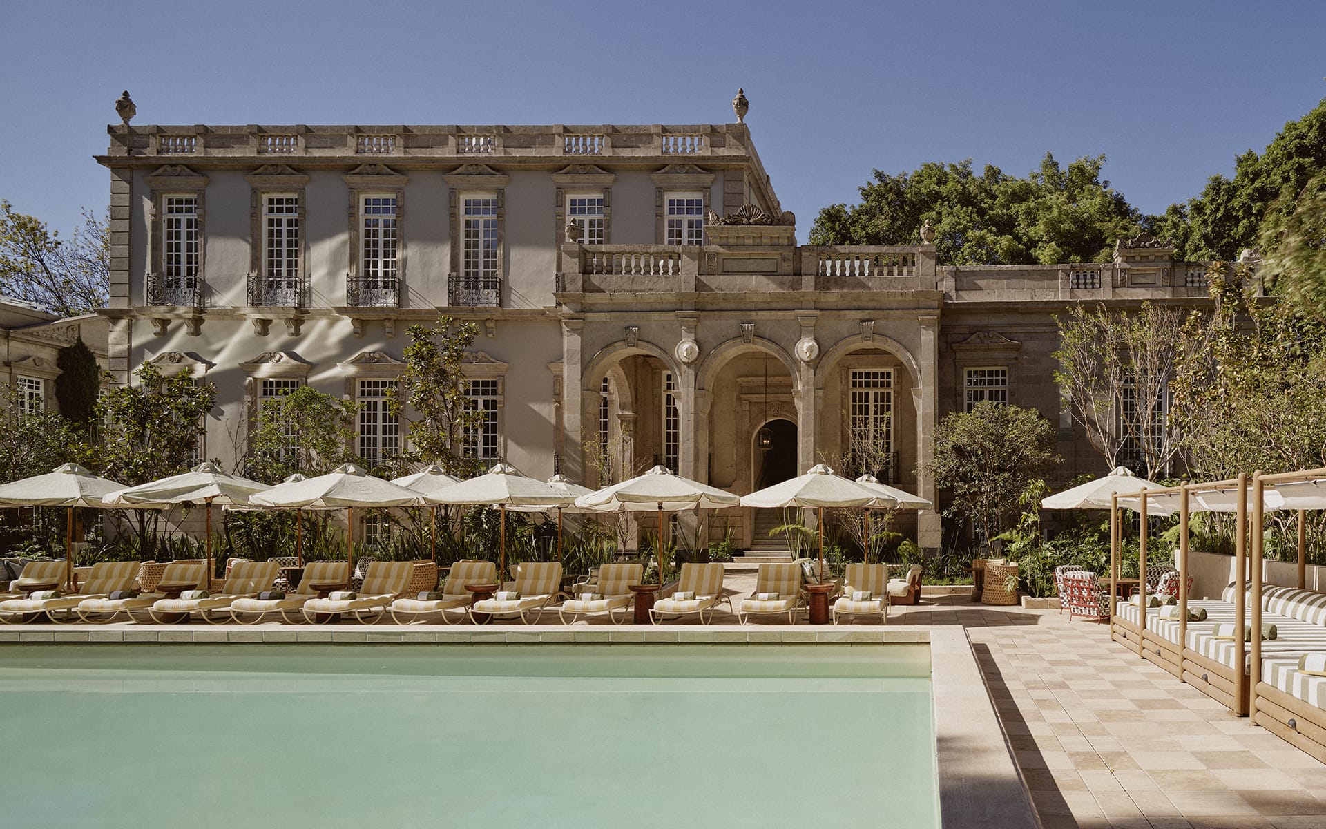 Luxury villa with elegant architecture, poolside lounge chairs, sun umbrellas, and surrounding greenery under a clear blue sky.
