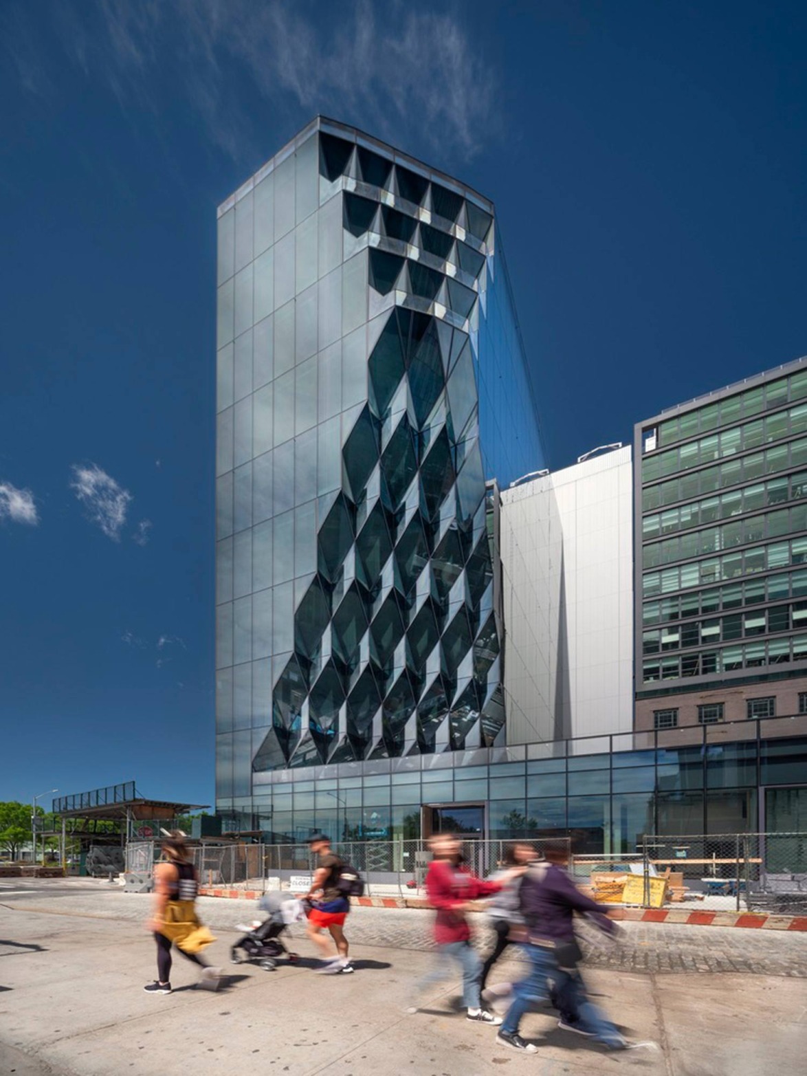 Modern glass building with geometric design, pedestrians walking by on a sunny day, construction zone in the foreground.