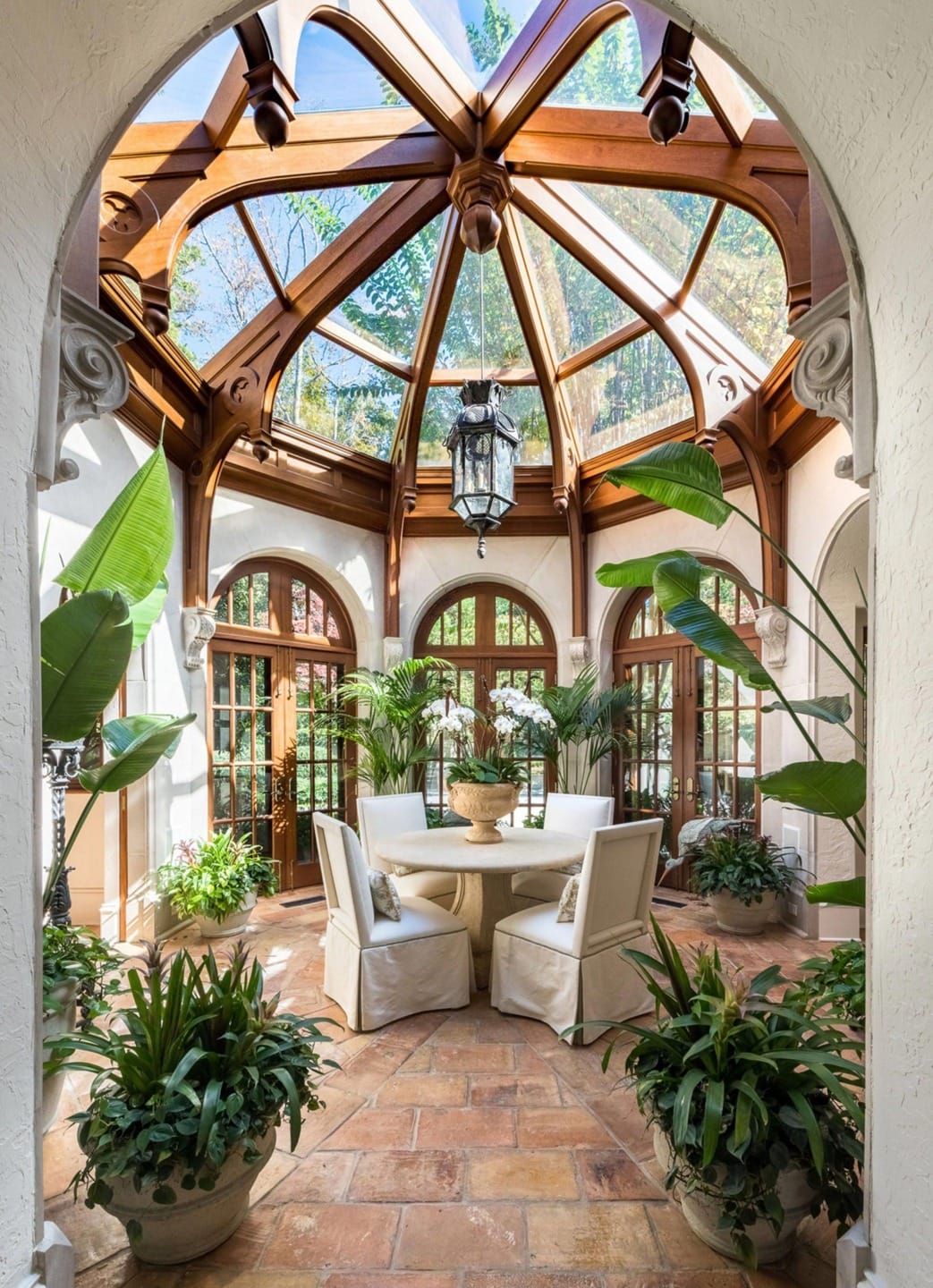 Sunlit conservatory with glass ceiling, wooden beams, round table, and lush plants surrounding a cozy seating area.