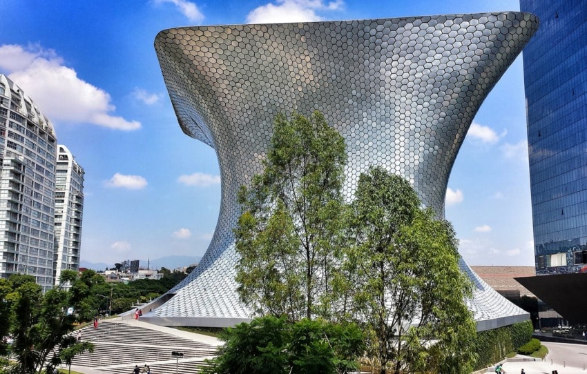 Futuristic silver museum building with a unique, curving design, surrounded by trees and modern cityscape.