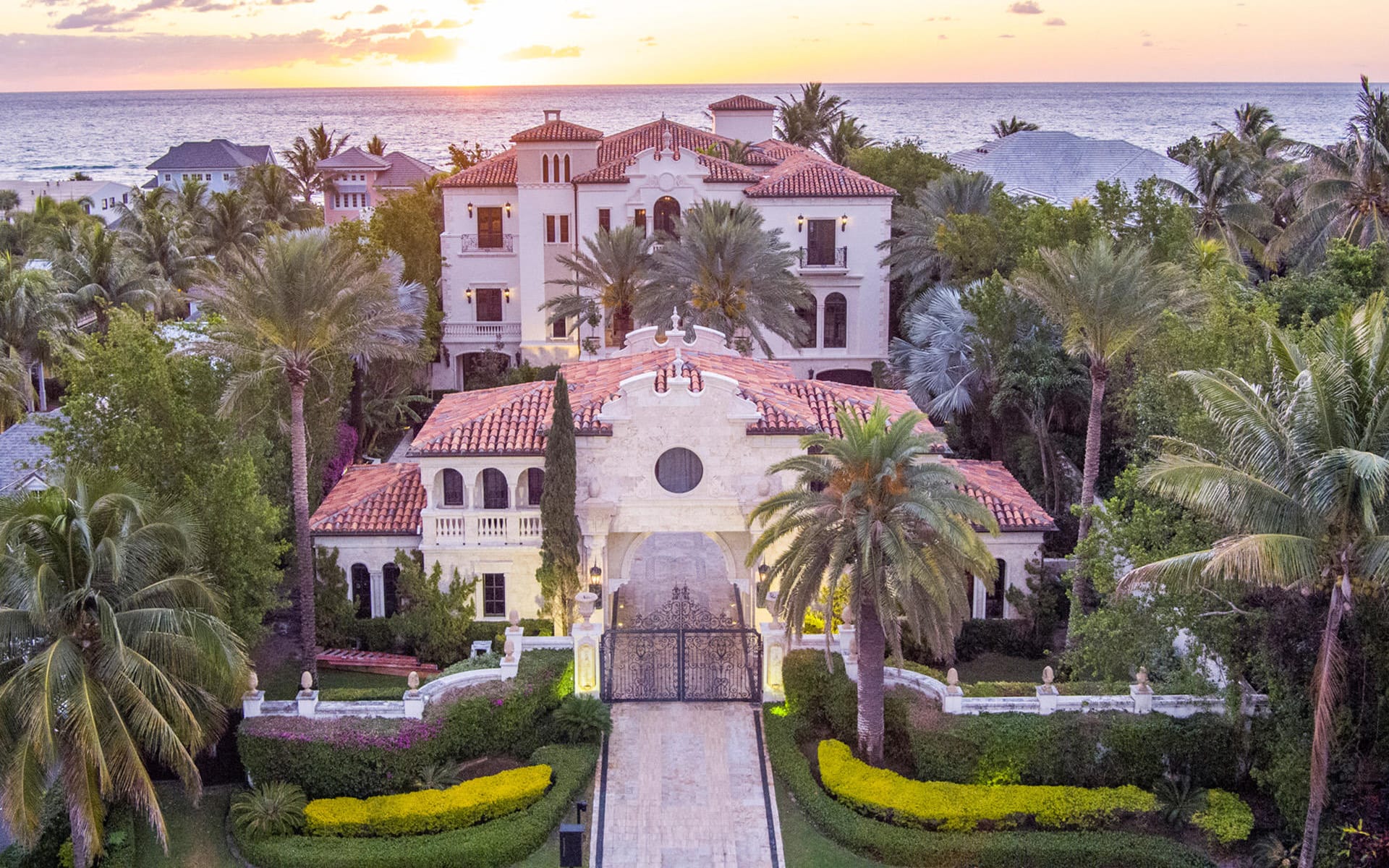 Luxurious mansion with red-tiled roofs, palm trees, and a sunset view over the ocean in an upscale neighborhood.