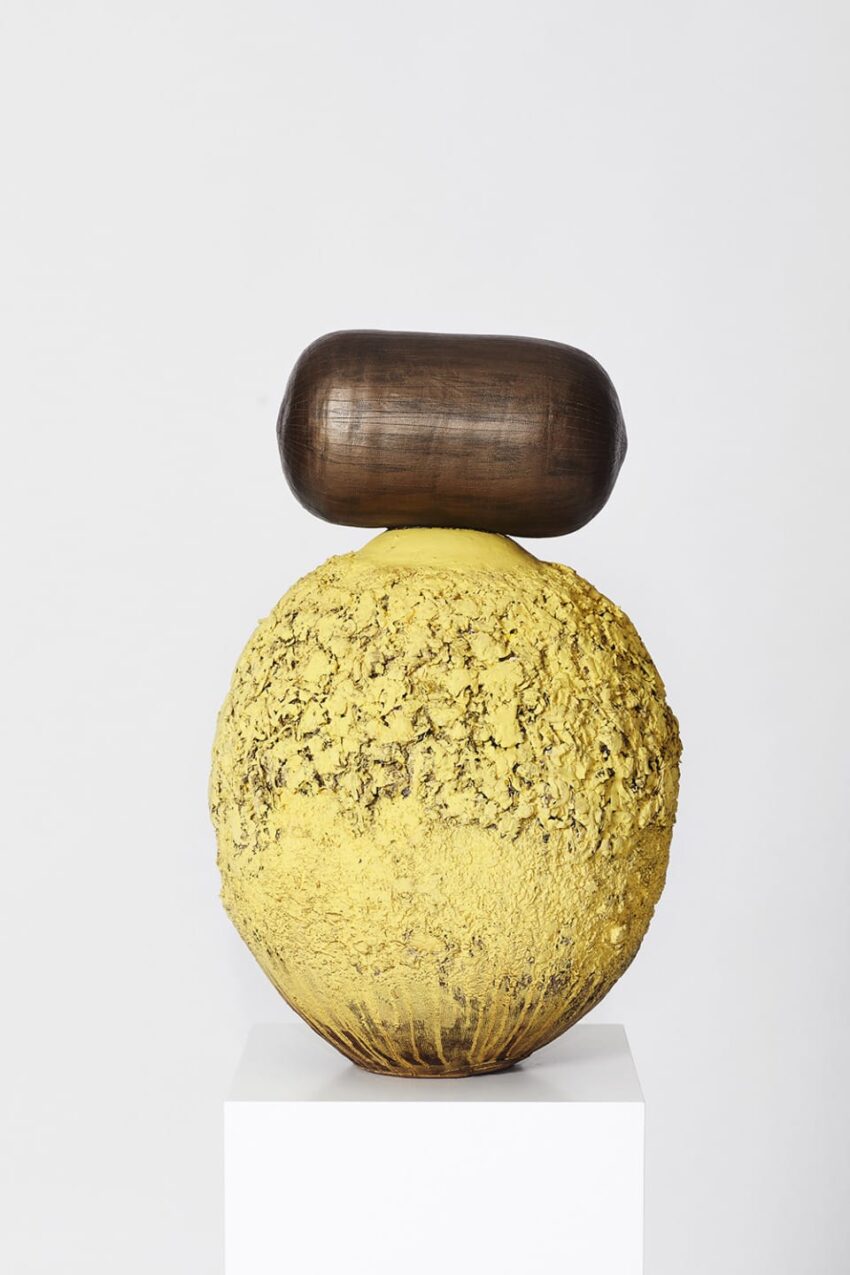 Abstract sculpture with a textured yellow sphere and smooth brown oblong shape on top, displayed on a white pedestal.