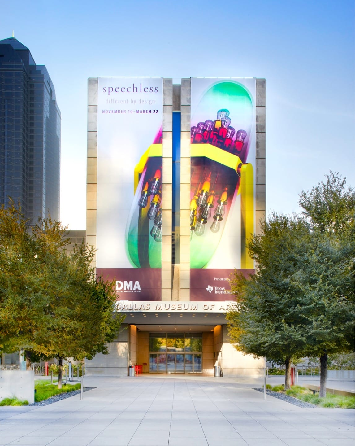 Museum entrance with large colorful banners promoting an exhibition titled "Speechless: Different by Design" amidst urban scenery.