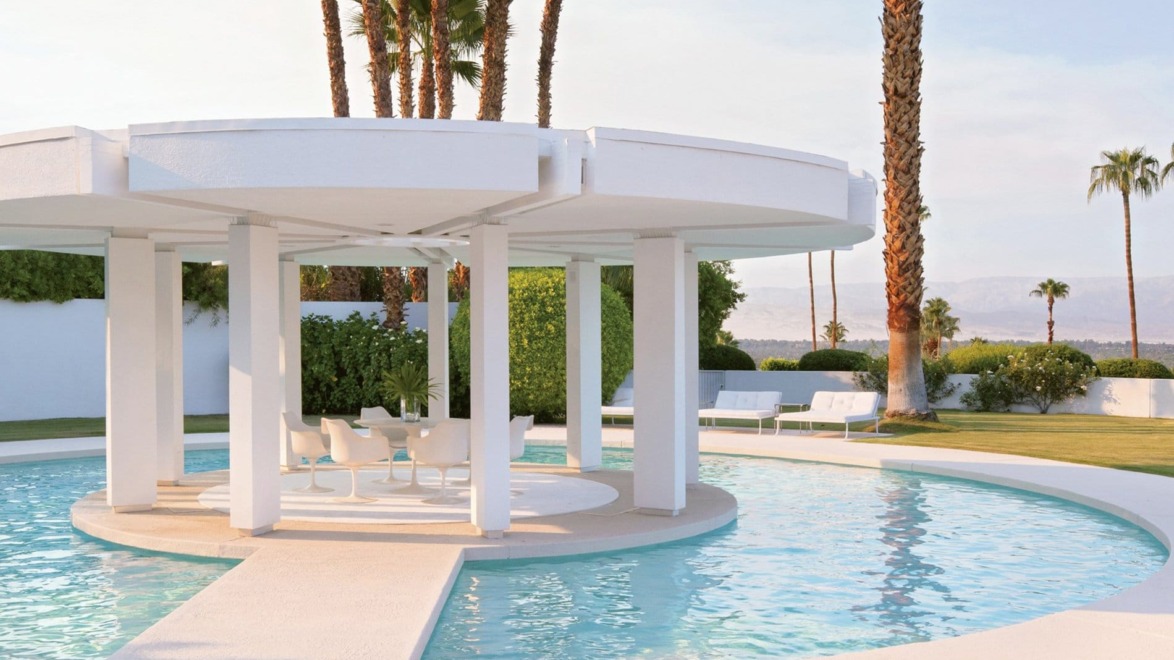 Outdoor patio with round canopy over pool surrounded by palm trees and modern white furniture in a sunlit garden setting.