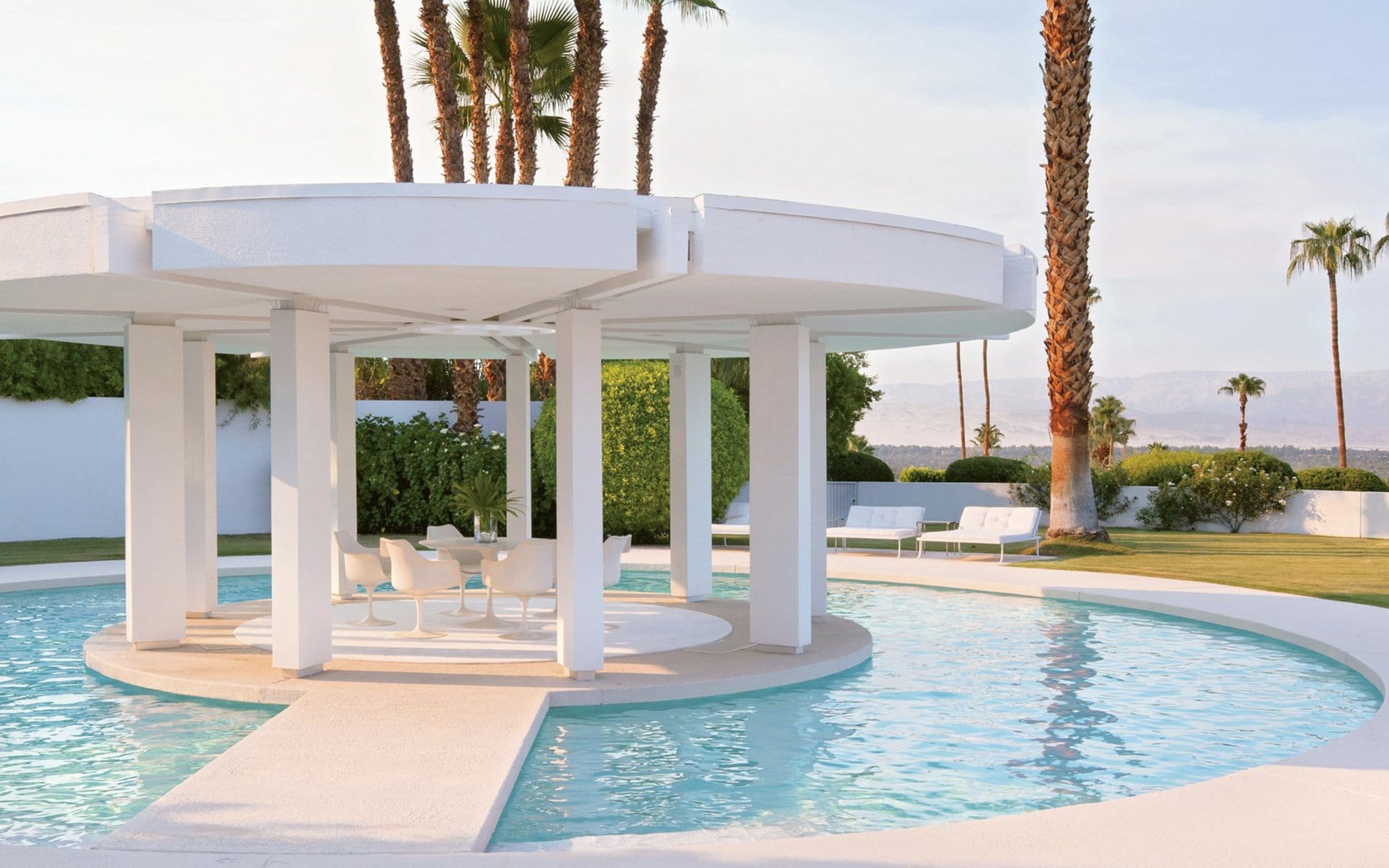 Outdoor patio with round canopy over pool surrounded by palm trees and modern white furniture in a sunlit garden setting.
