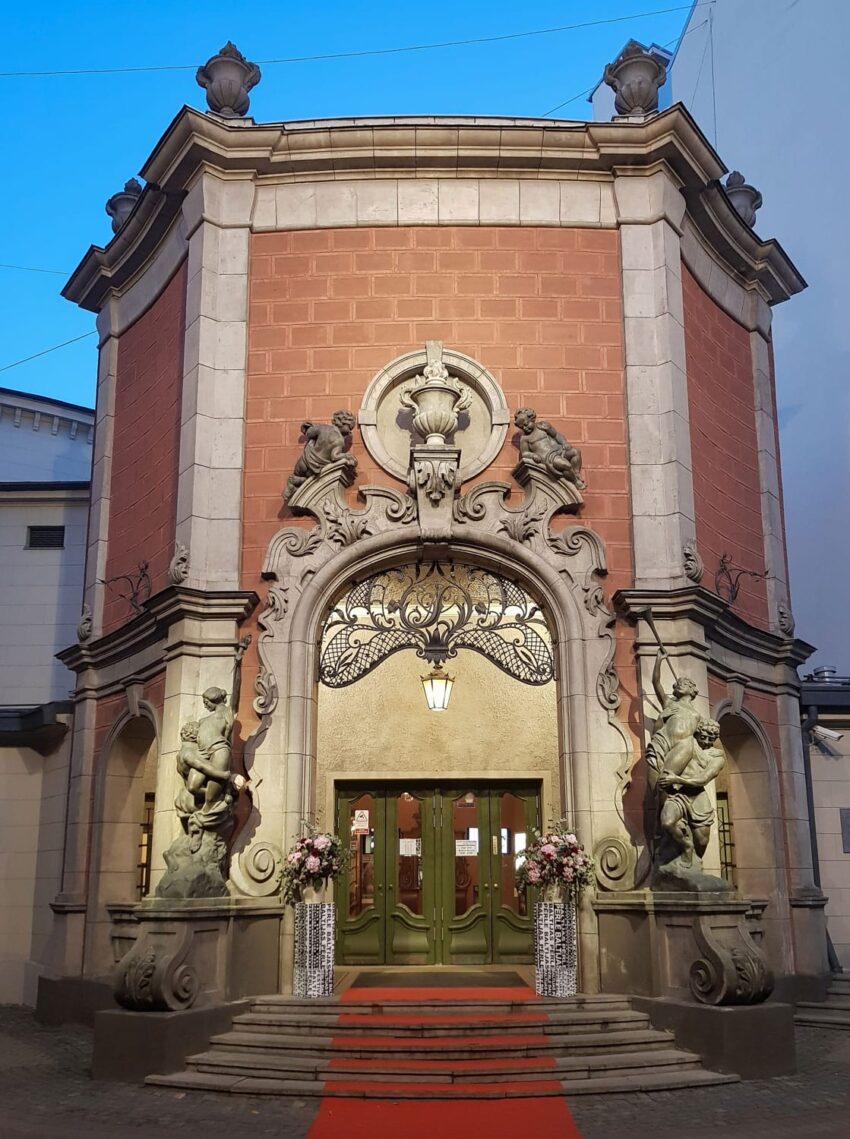 Ornate building entrance with decorative statues, red brick façade, floral arrangements, and a red carpet leading to the door.