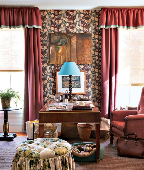 Home office with floral wallpaper, red curtains, a wooden desk, blue lamp, and cozy chair with plant accents.