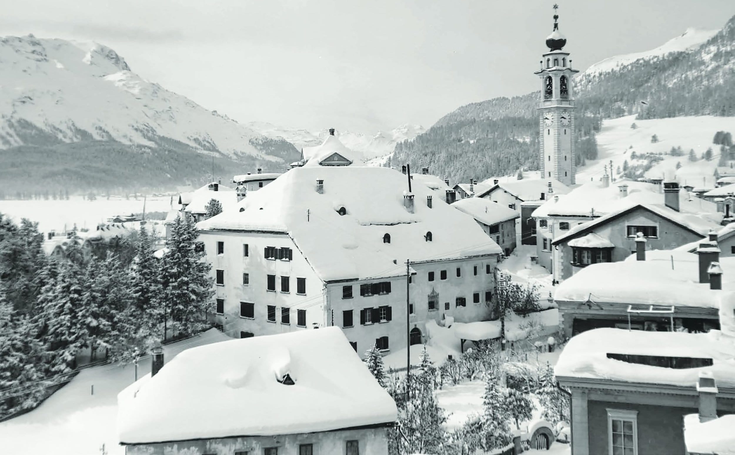 Snow-covered village in the mountains with traditional buildings and a clock tower under a cloudy sky.