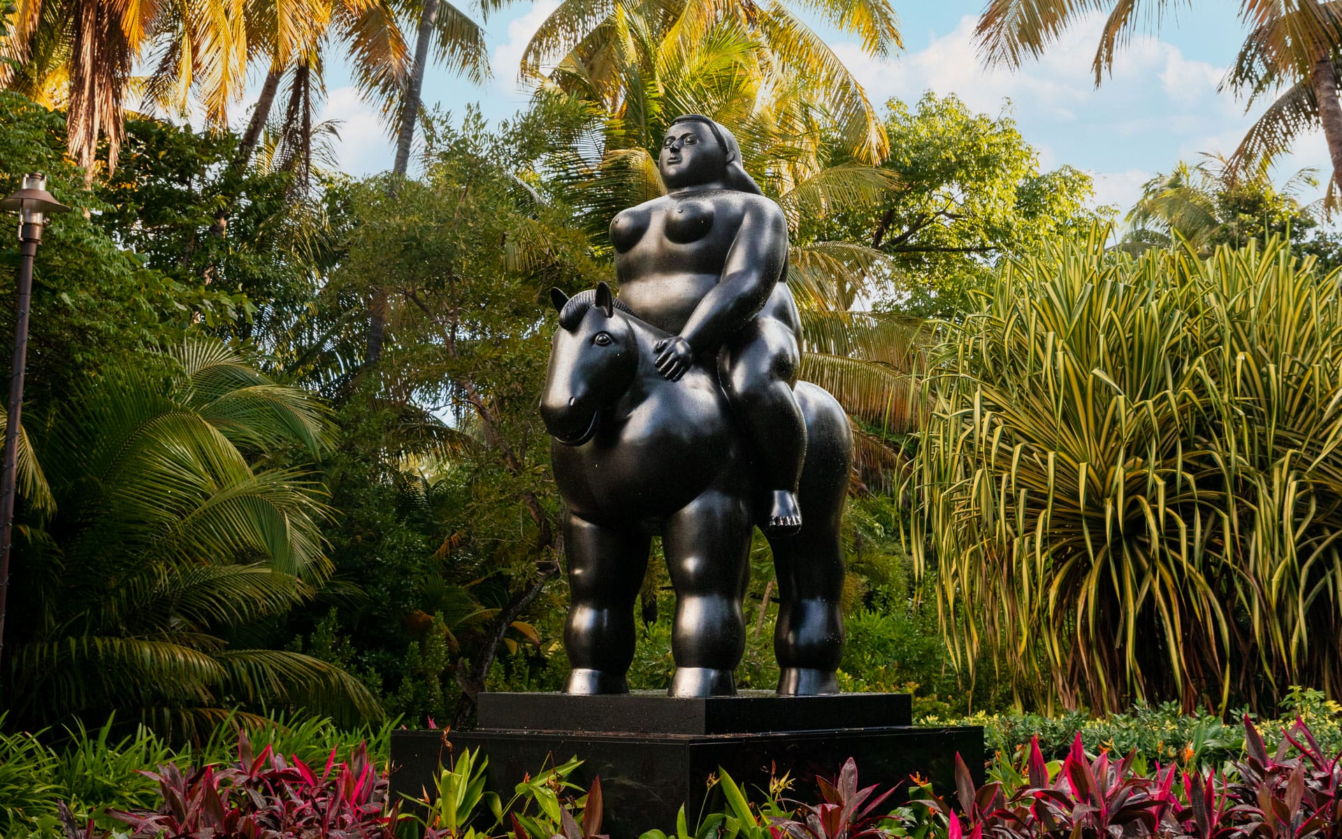 Statue of a rotund figure riding a horse amid lush green tropical plants and palm trees in a garden setting.