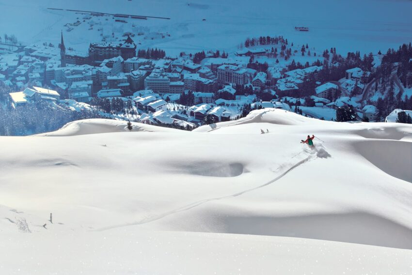 Skier carving through fresh snow on a sunny day with snowy mountain village in the background.