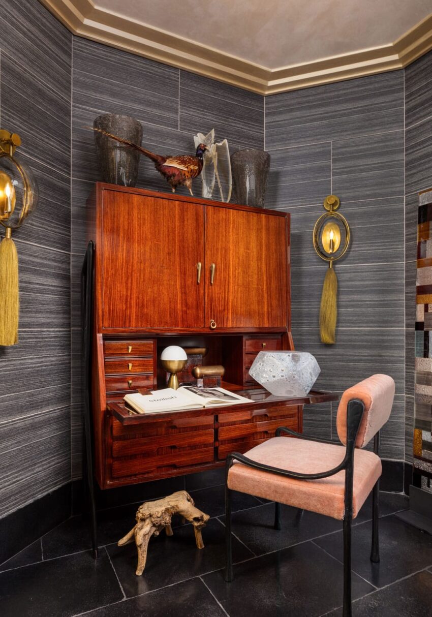 Mid-century modern desk with open drawers, decorative items, and a pink upholstered chair in a stylish, dark-paneled room.