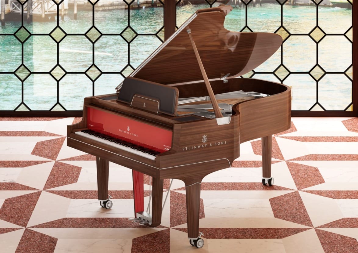 Grand piano with open lid in a bright room with geometric floor tiles and large windows overlooking a water view.