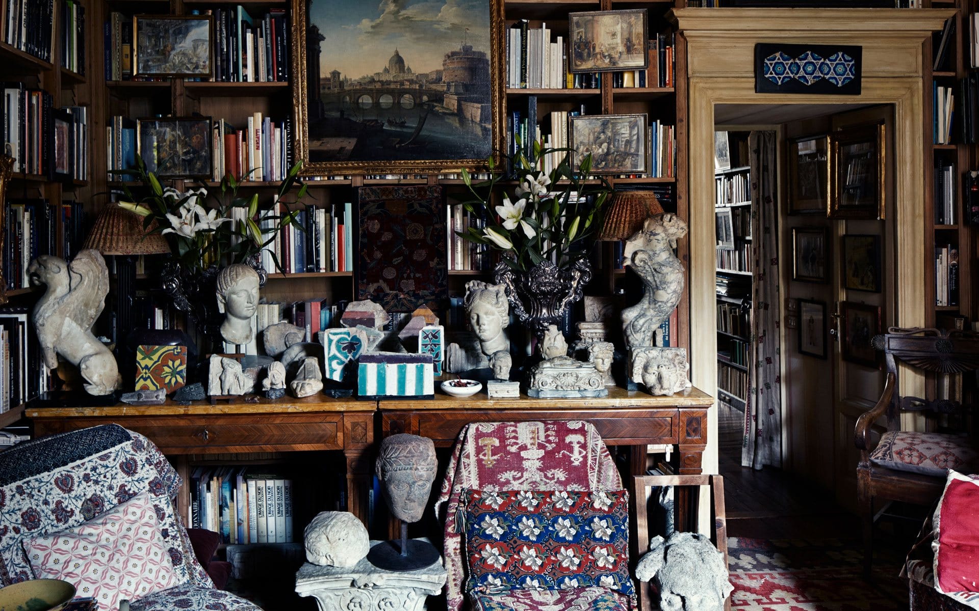 Eclectic library interior with books, sculptures, and paintings on shelves and tables, creating a cozy and artistic atmosphere.