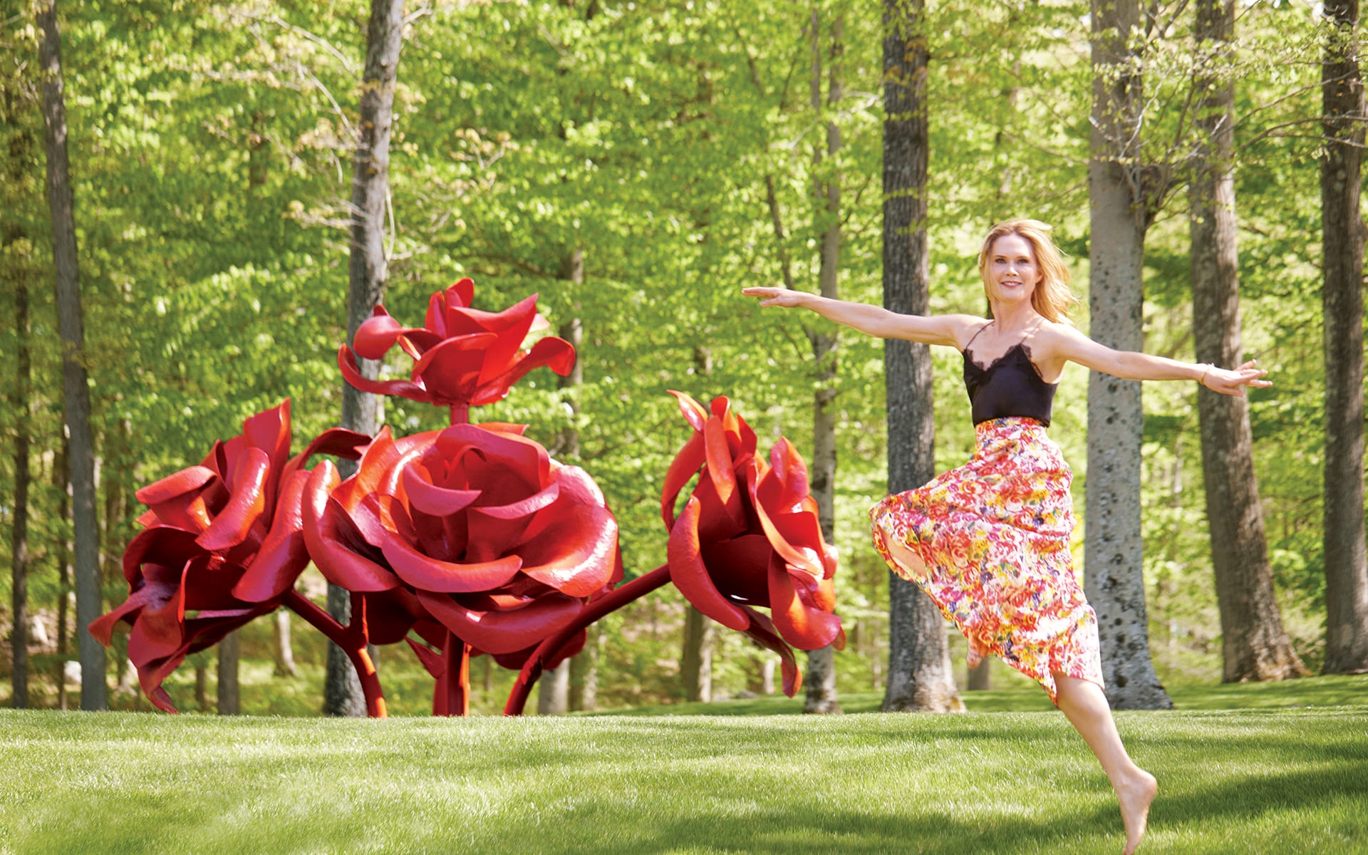 Woman dancing joyfully on grass near large red rose sculptures in a sunlit forest setting.