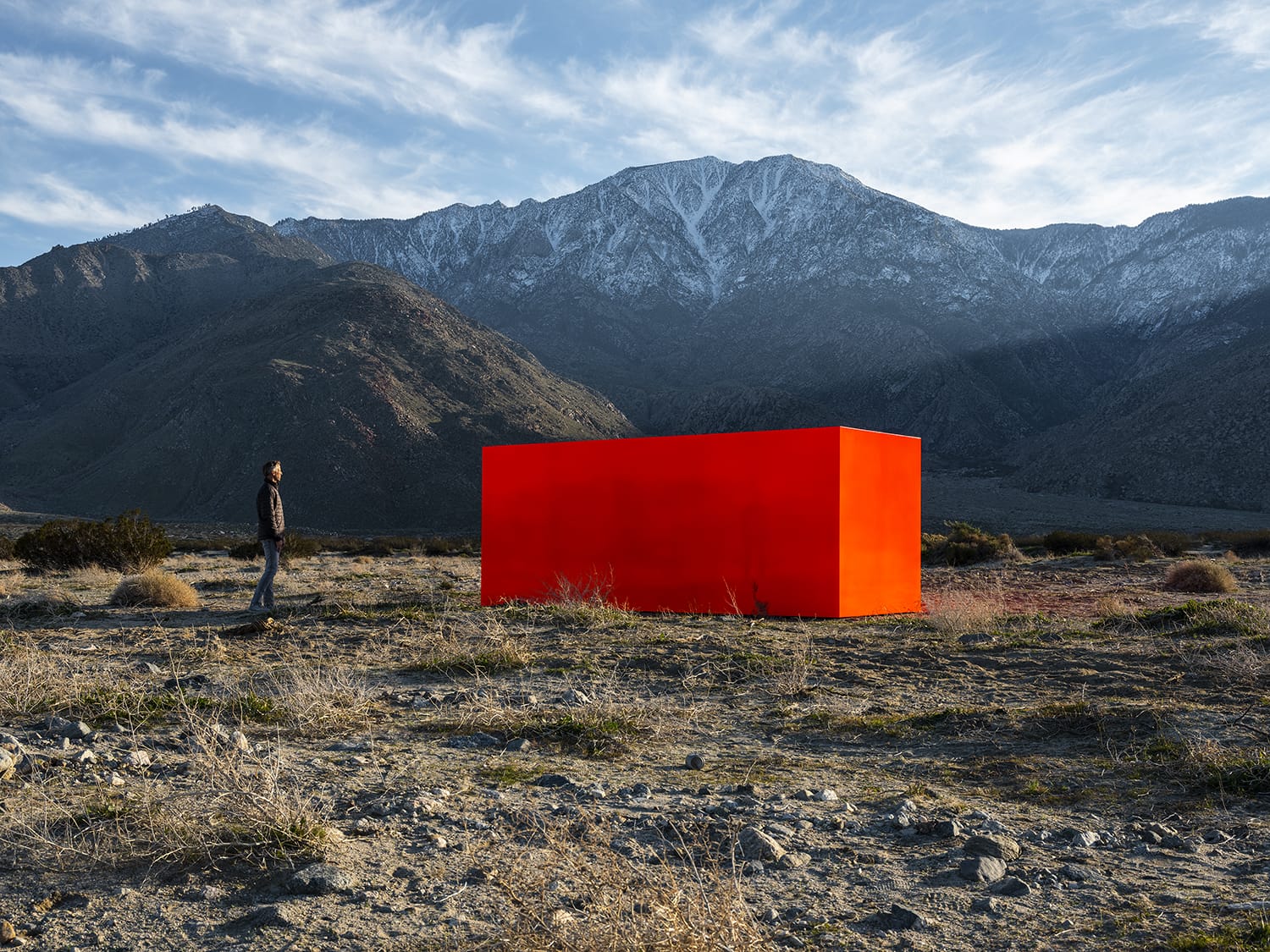 Person standing in a rocky desert landscape, gazing at a large vibrant red rectangular structure with mountains in the background.