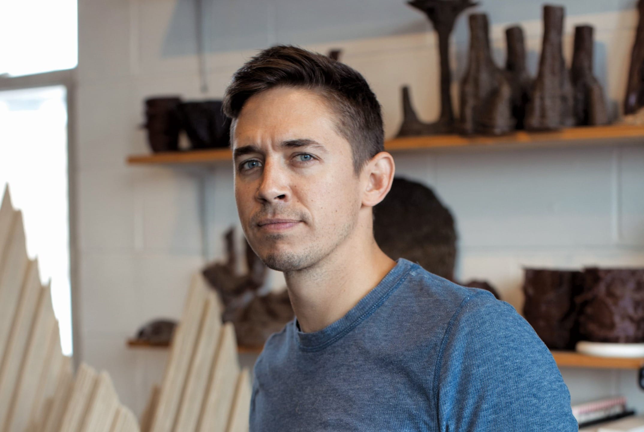 Man in a blue shirt standing in a room with shelves filled with artistic pottery pieces in the background.