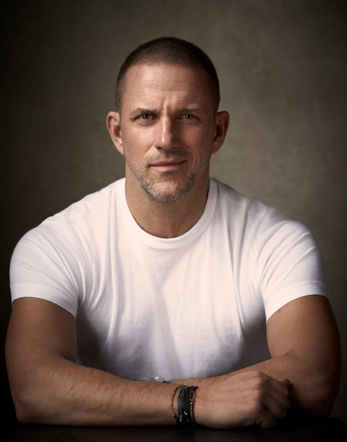 Man in a white t-shirt sitting with arms crossed on a table, looking directly at the camera.