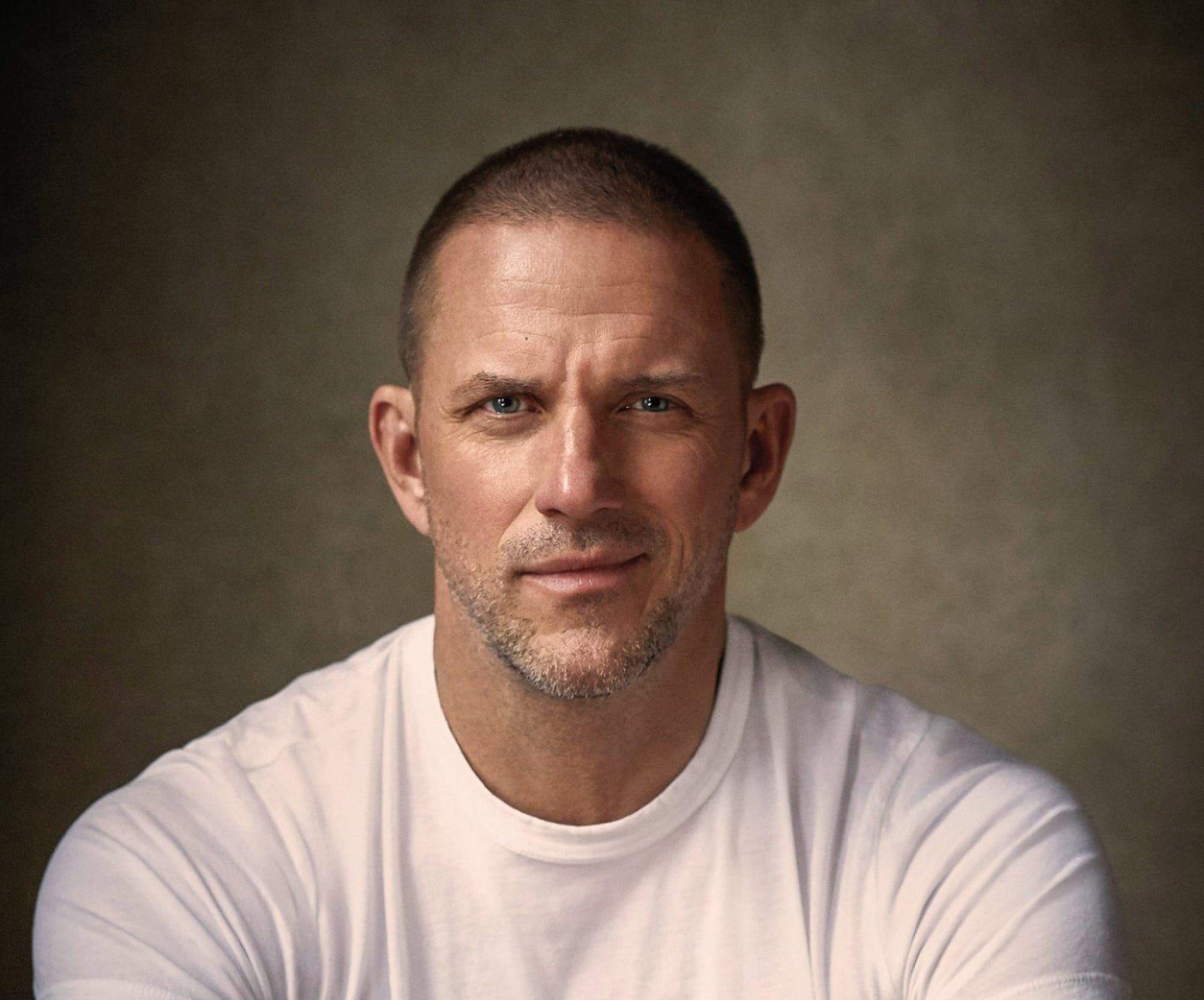 Man with a beard and short hair wearing a white T-shirt against a neutral background, looking directly at the camera.
