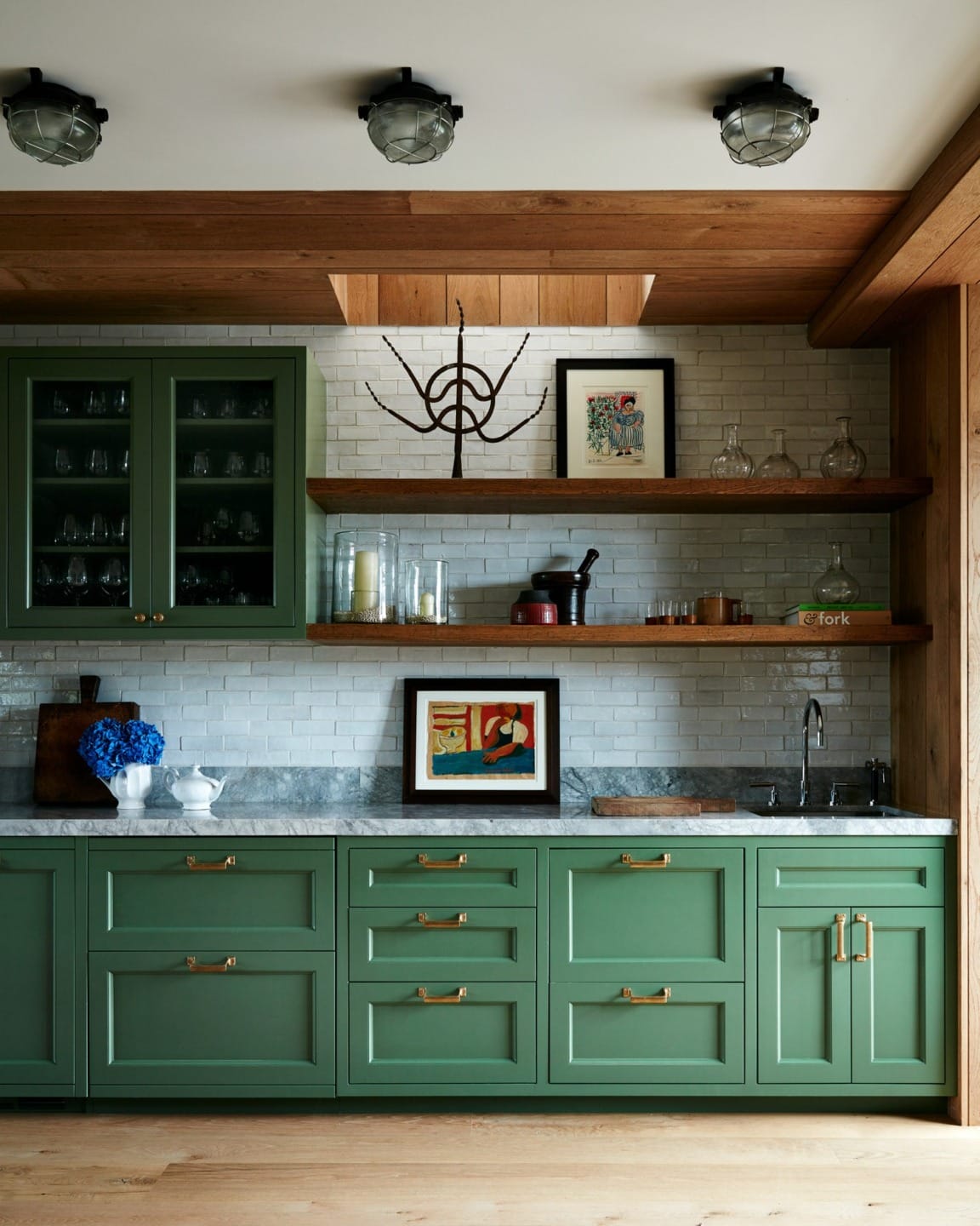 Modern kitchen with green cabinets, marble countertop, wooden shelves, artwork, and stainless steel fixtures.