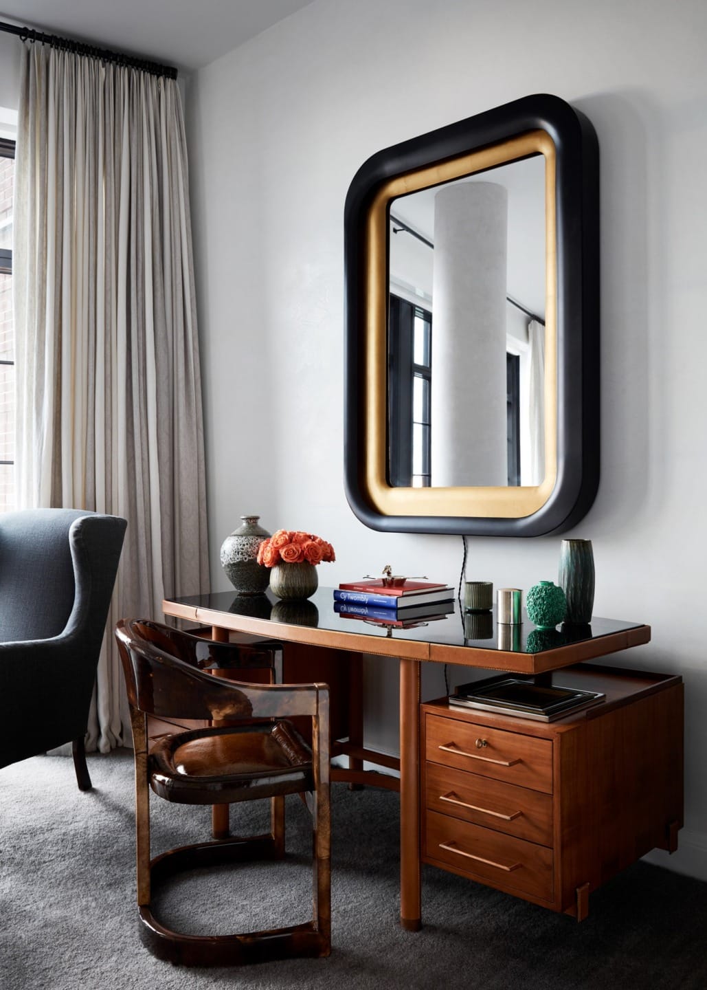 Stylish home office with wooden desk, modern chair, decorative vases, books, and large mirror on a white wall.