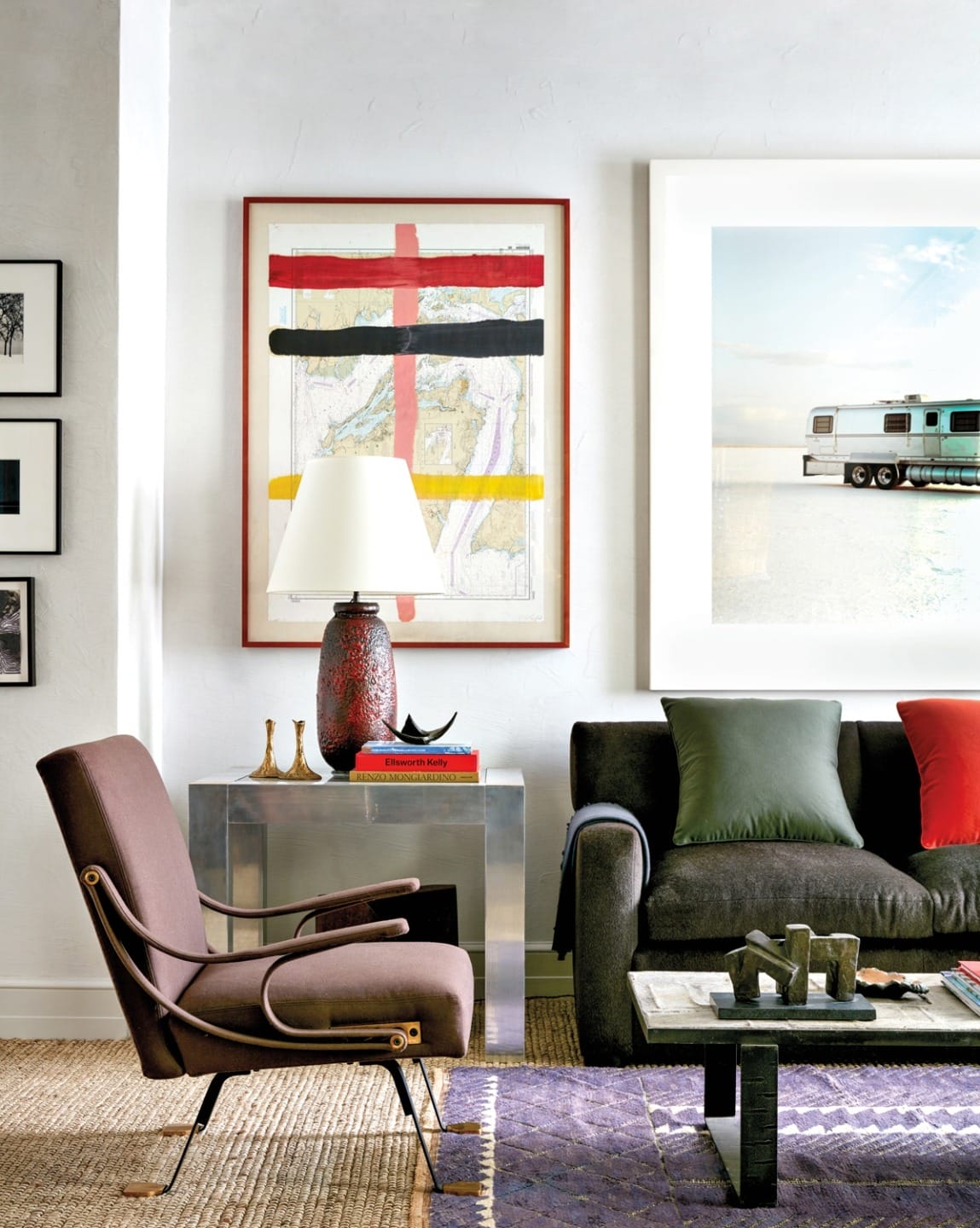 Modern living room with brown chair, glass side table, red lamp, abstract wall art, dark sofa, and colorful pillows.