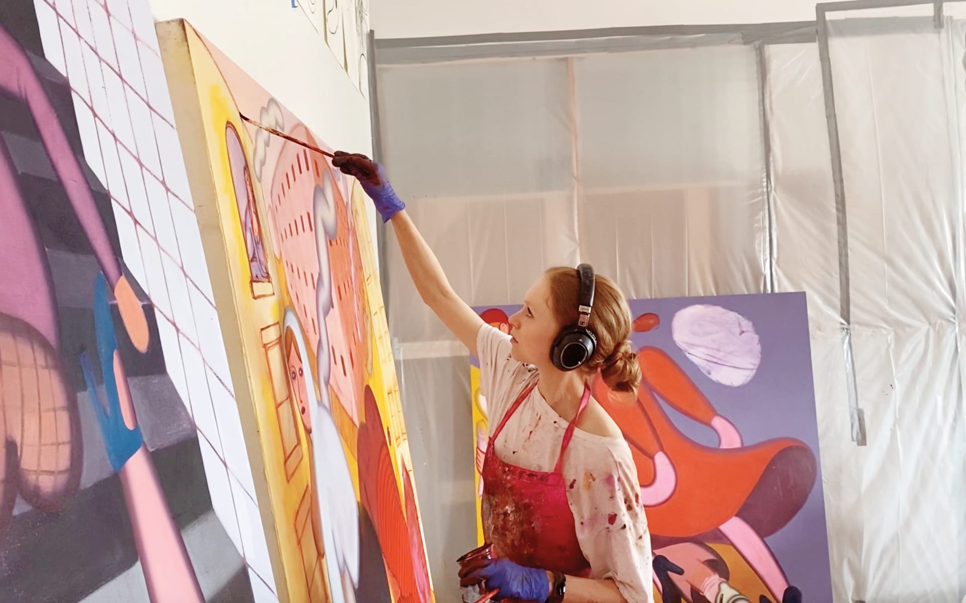Artist in headphones paints a large abstract mural, wearing paint-splattered apron and gloves in a bright studio.