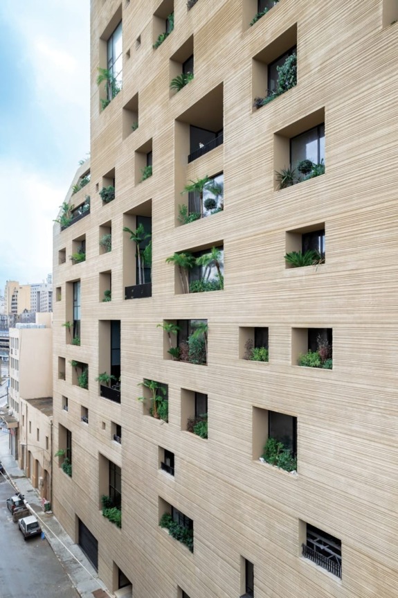 Modern beige building with multiple square windows and green plants, exterior view on an urban street.
