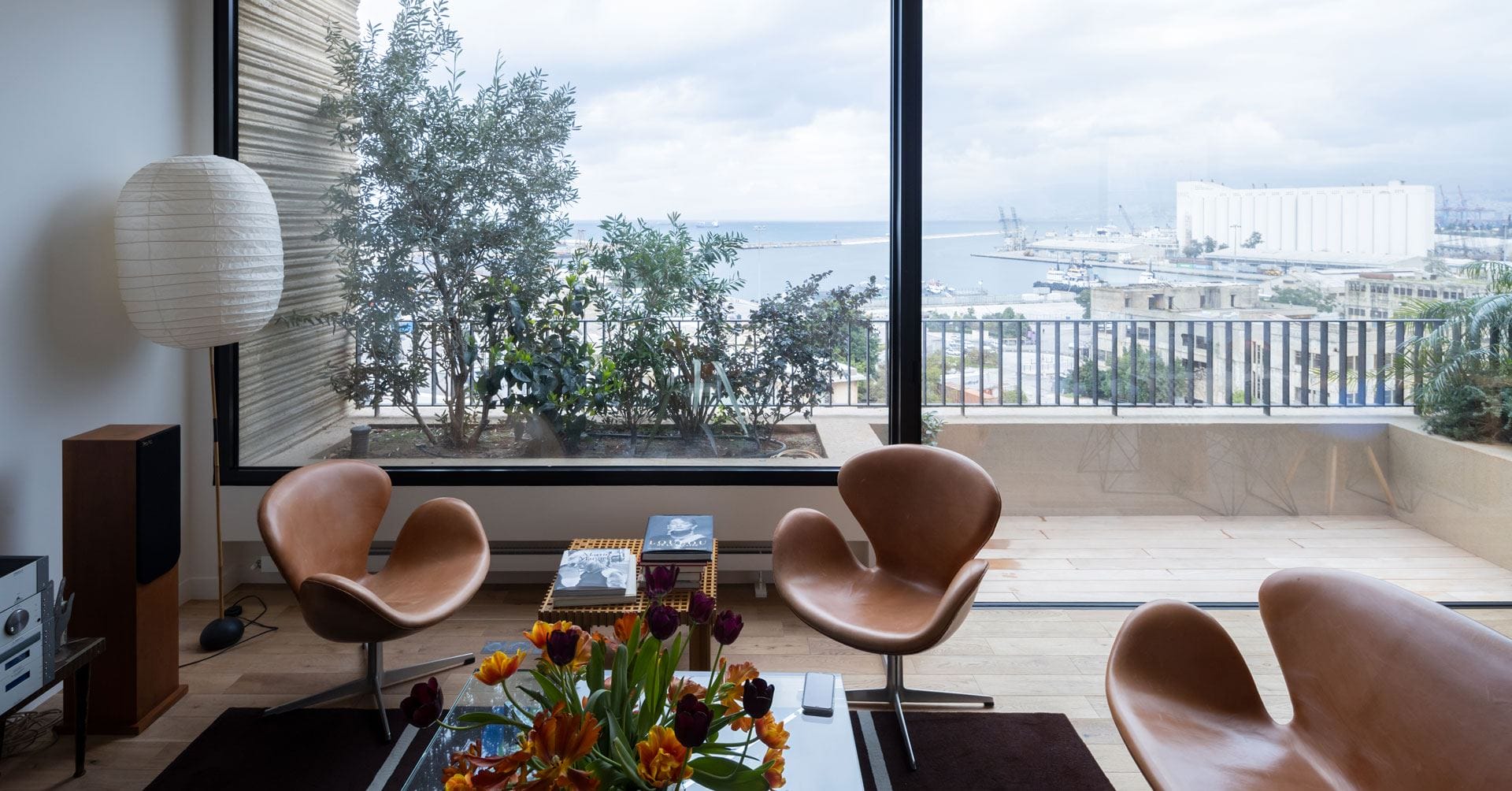 Modern living room with tan chairs, a coffee table with flowers, and a large window overlooking a coastal view with ships.
