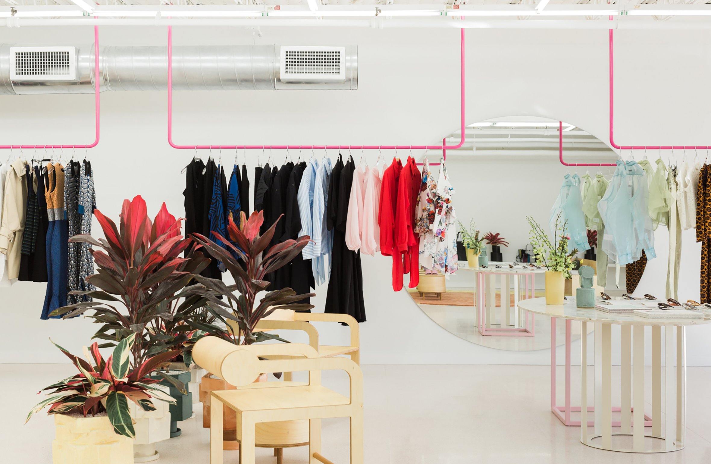 Modern clothing boutique with colorful dresses on racks, vibrant plants, wooden chairs, and a large circular mirror.