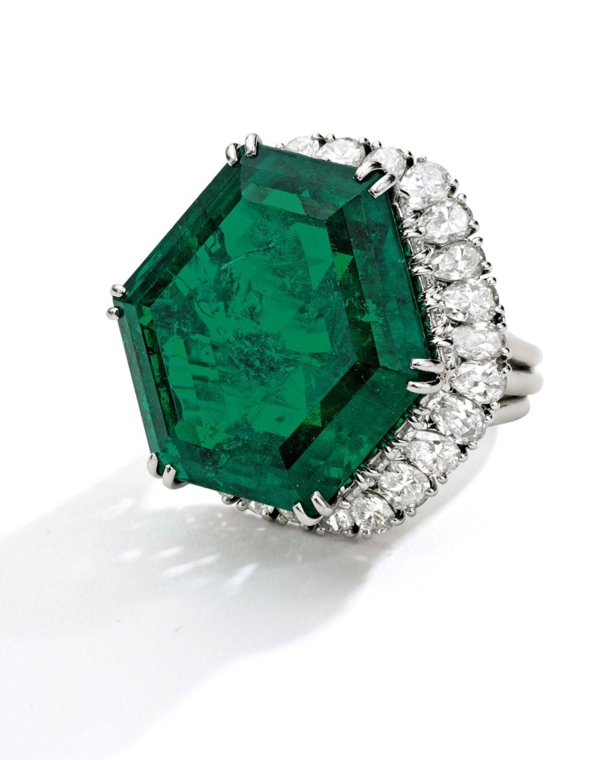 Hexagonal emerald ring with a halo of diamonds, set in white gold, on a reflective surface.