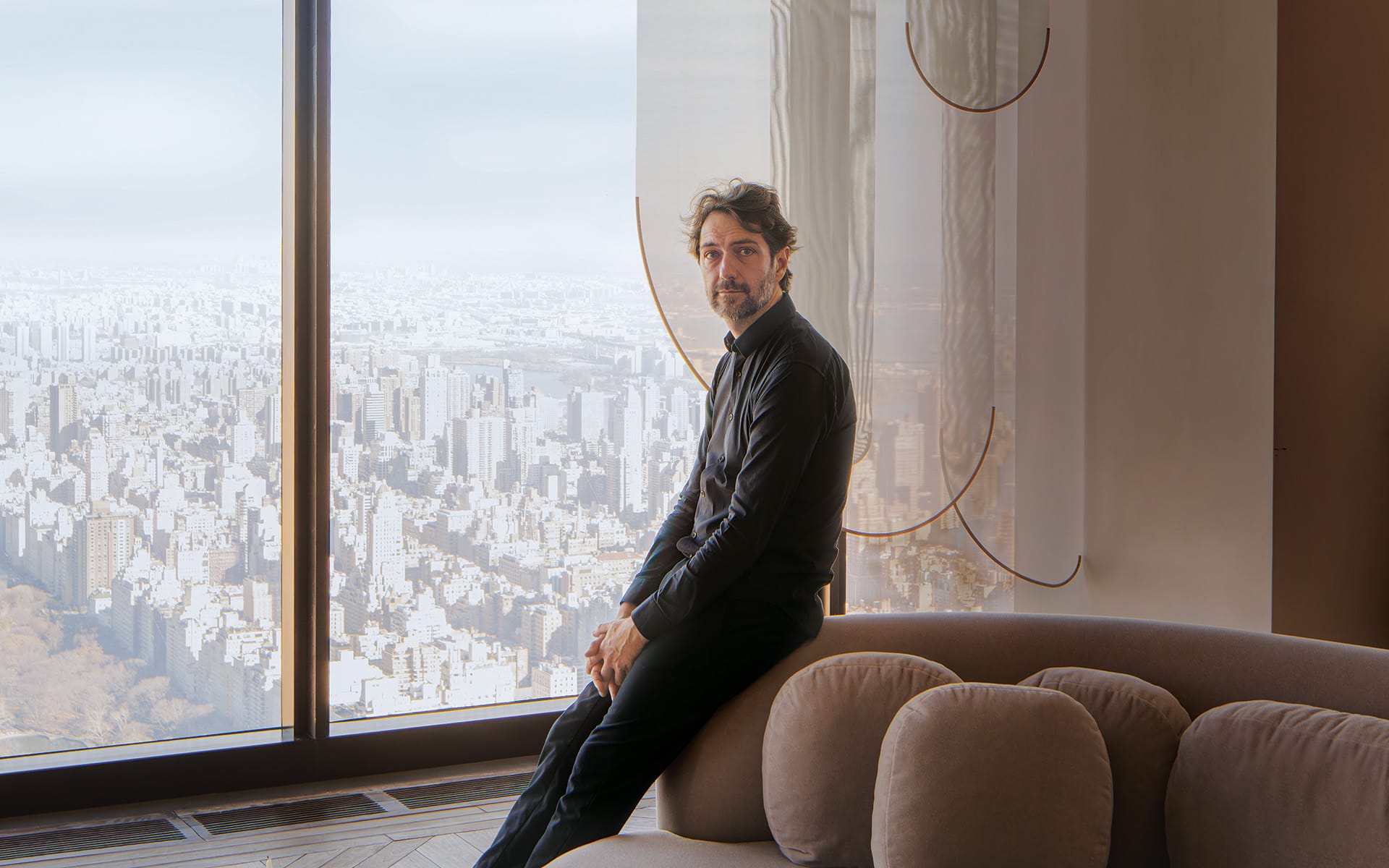 Man sitting on a sofa by a large window with a cityscape view in the background