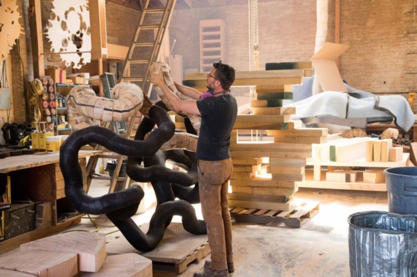 Artist working on large sculptural piece in a cluttered woodshop filled with tools, wood, and unfinished projects.