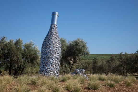 Sculpture of a large wine bottle made from metallic scraps surrounded by grass and olive trees against a clear blue sky.