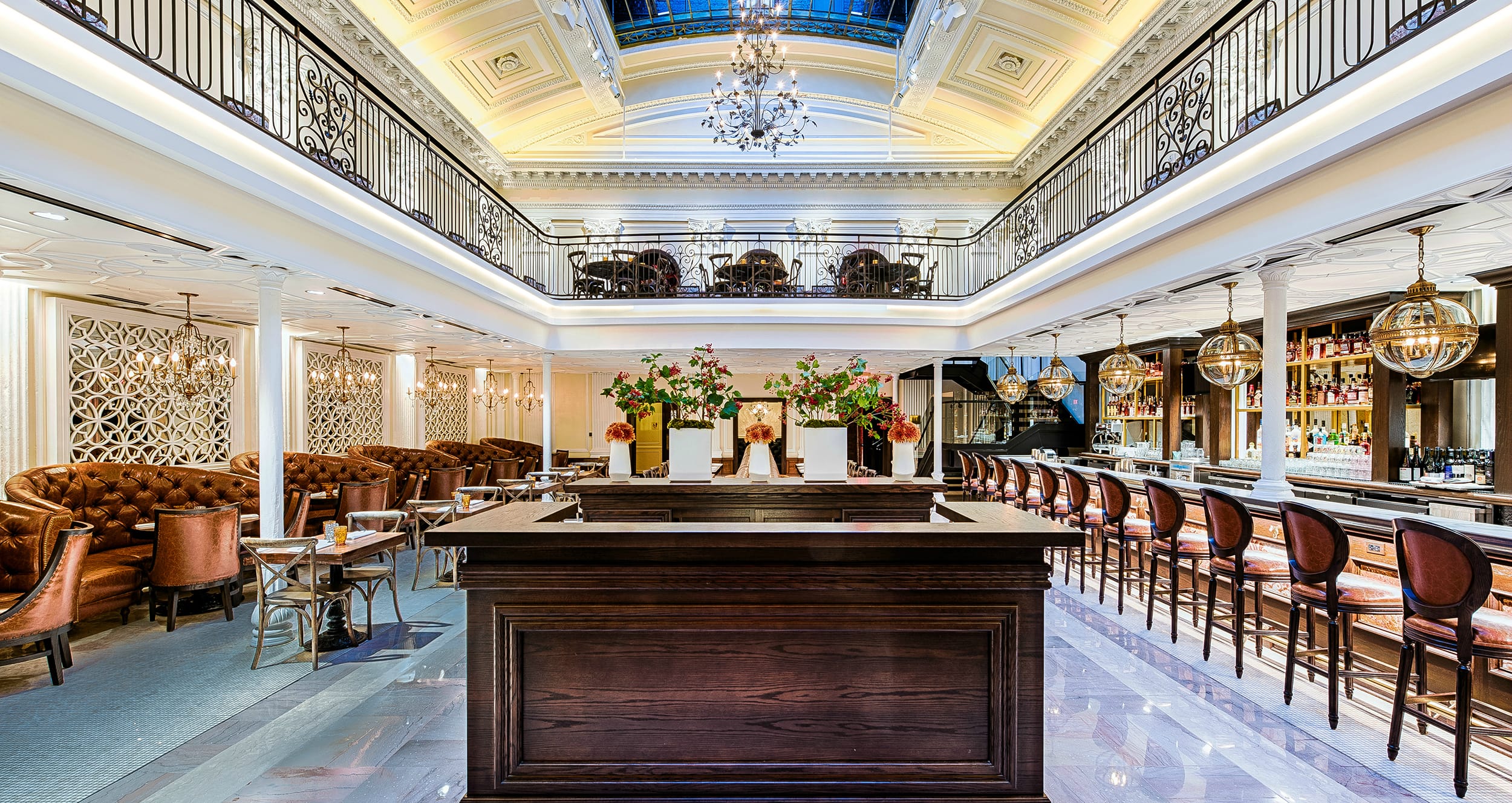 Elegant, spacious restaurant interior with high ceilings, chandeliers, bar counter, and leather seating with floral arrangements.
