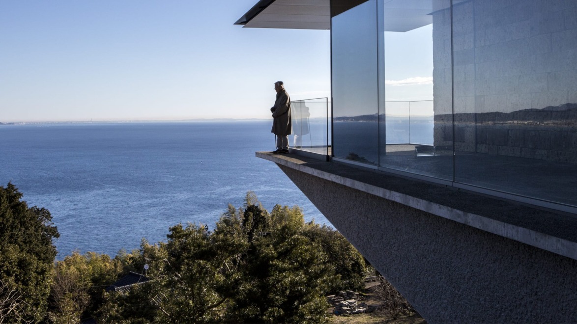 Person standing on a modern building's balcony overlooking a vast ocean and landscape on a clear day.