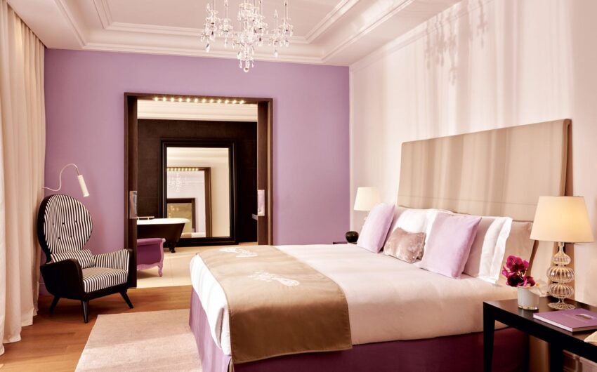Luxurious hotel room with lavender walls, elegant bed, armchair, chandelier, and large mirror reflecting a cozy ambiance.