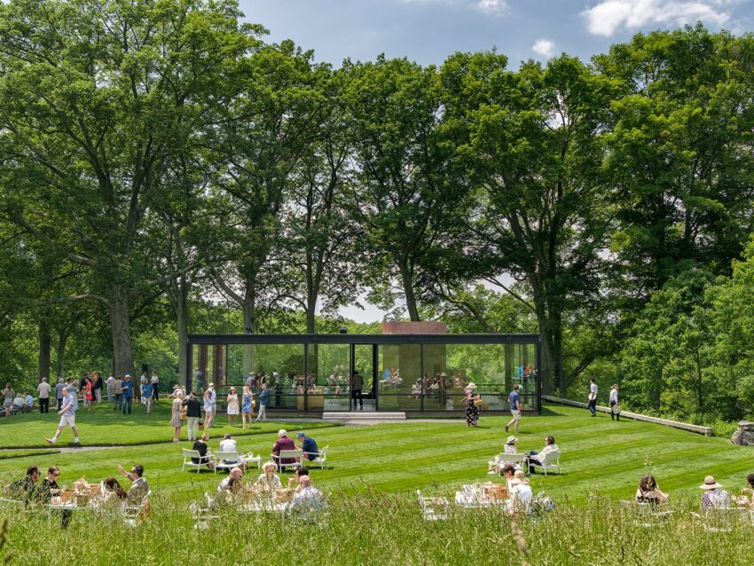People enjoying an outdoor event on a sunny day near a modern glass building surrounded by trees and greenery.