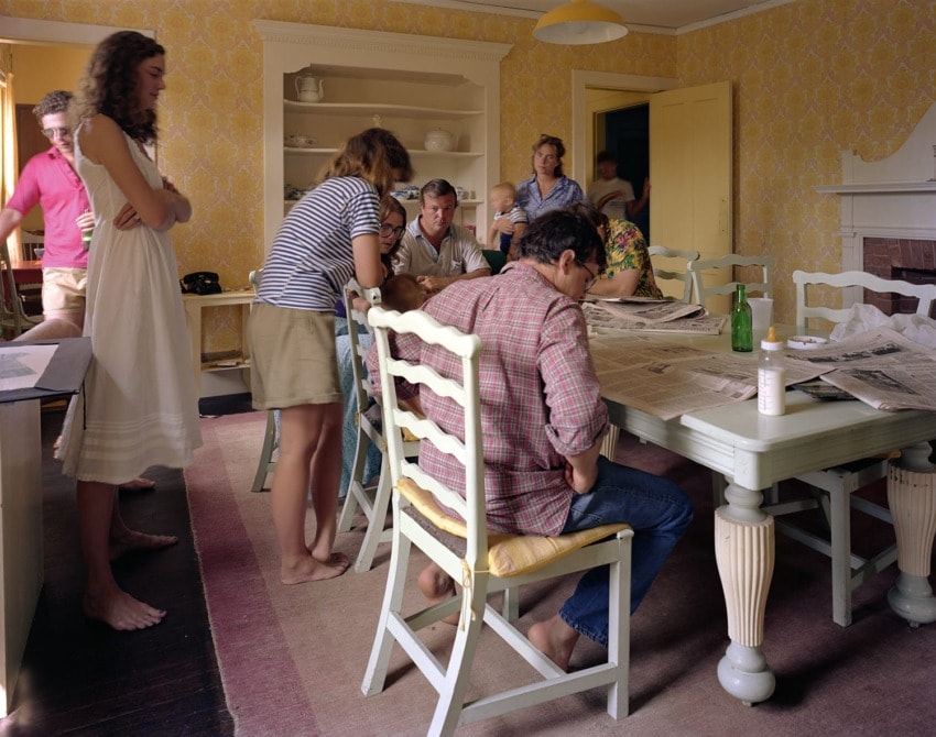 People gathered around a dining table reading newspapers in a vintage-style room with yellow wallpaper.