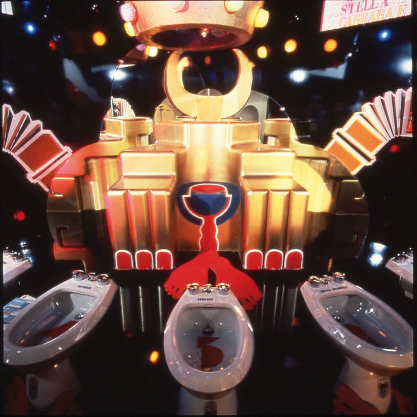 Colorful, surreal art installation featuring crowns, geometric shapes, and three white bidets with reflections.