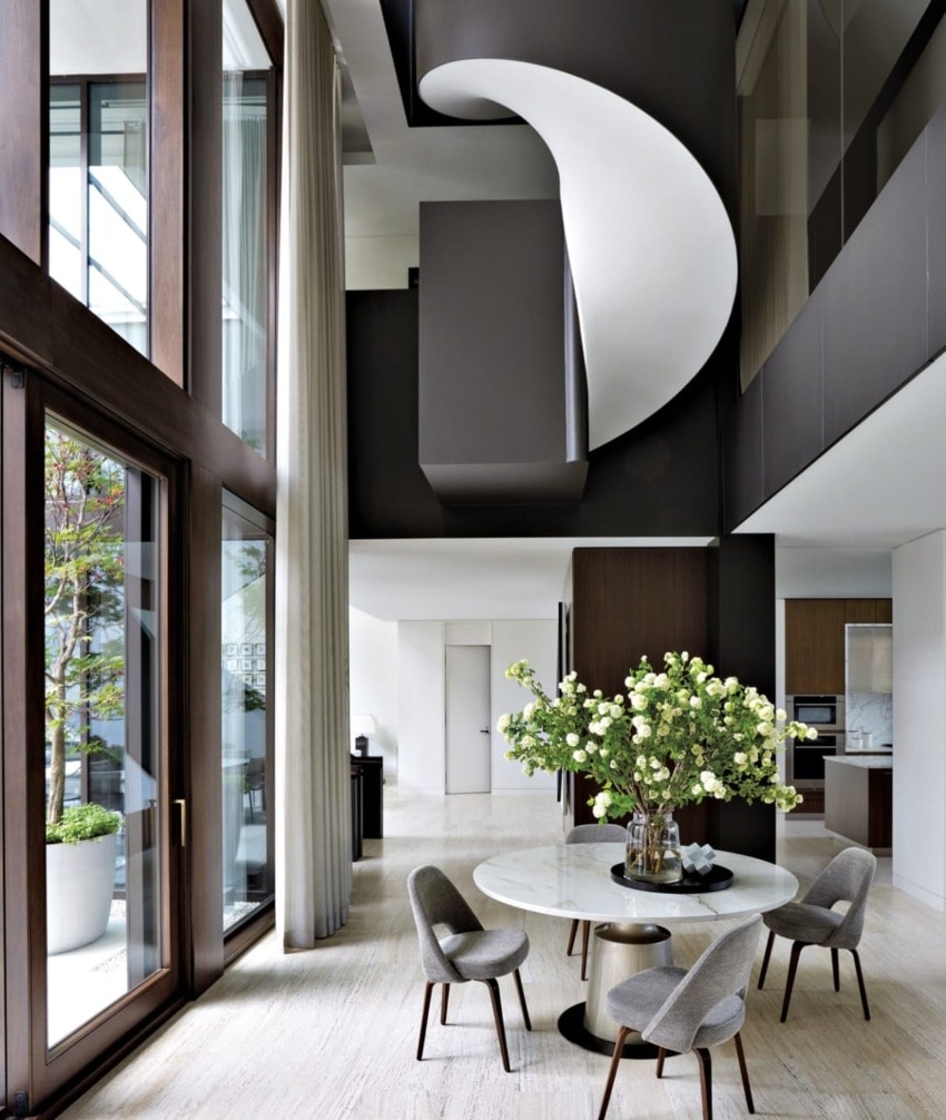 Modern open-plan living area with high ceilings, a spiral staircase, large windows, a table with four chairs and floral centerpiece.