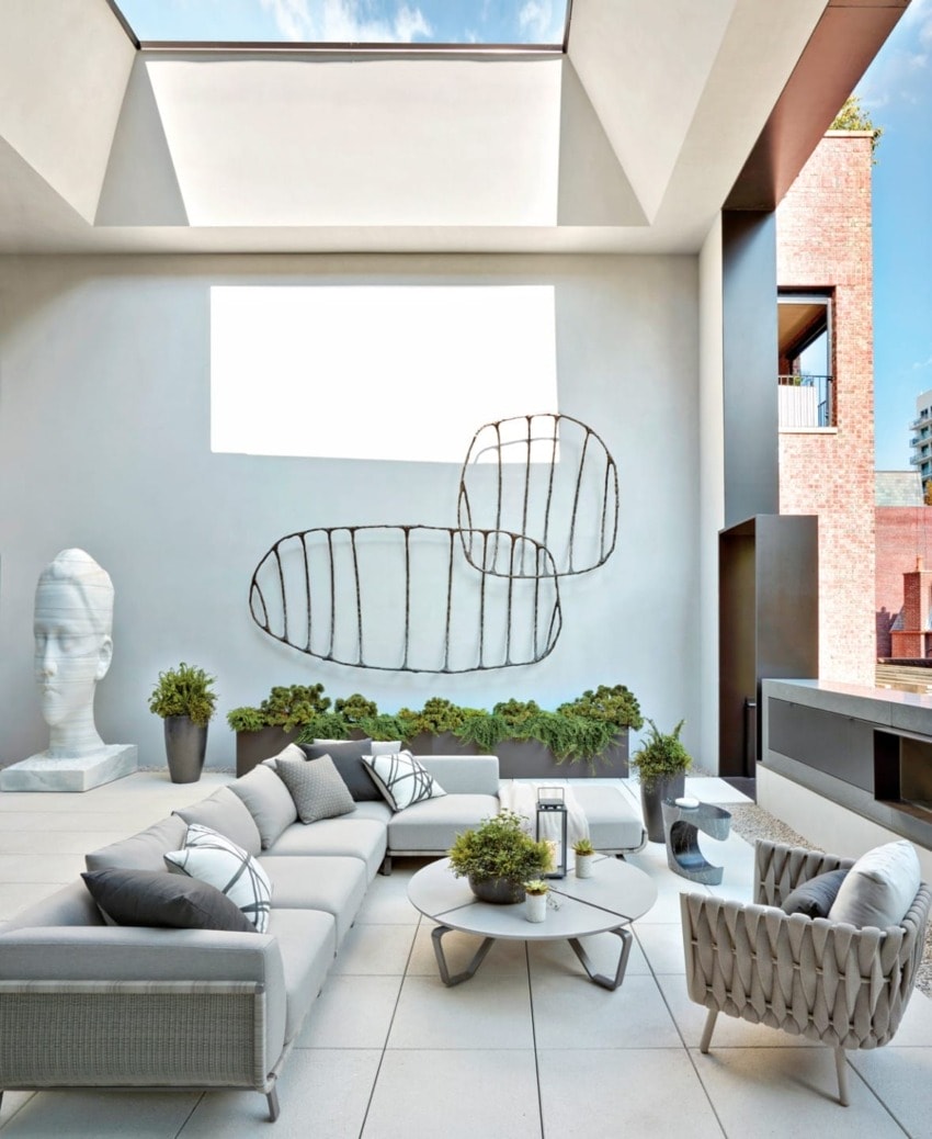 Modern outdoor terrace with skylight, contemporary furniture, abstract wall art, and potted plants on a sunny day.