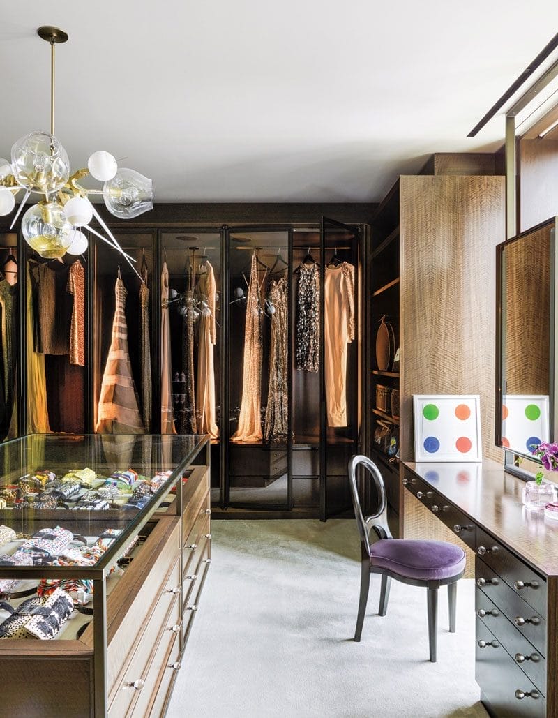 Luxurious walk-in closet with elegant dresses, modern chandelier, glass display drawers, and a dressing table with a chair.