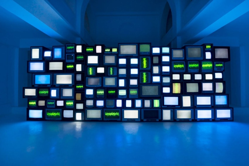 Wall of vintage televisions with varied static patterns and blue ambient lighting in a dimly lit room.
