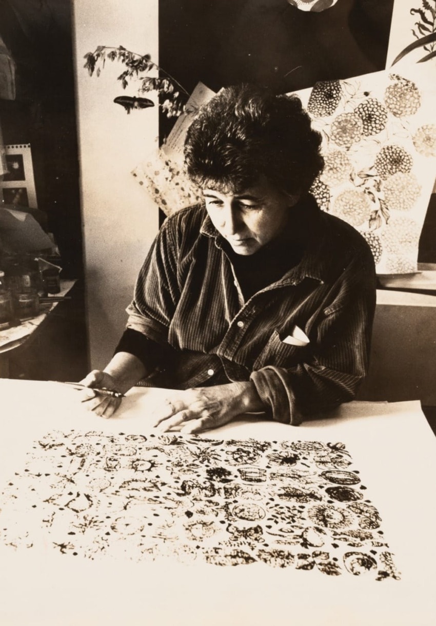 Artist working on a detailed patterned design at a desk, surrounded by artwork and plants.