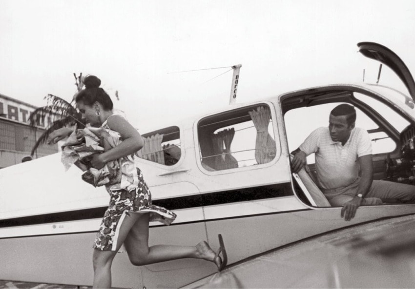 Woman in dress hurries away from small airplane while man watches from open door, airport background visible.