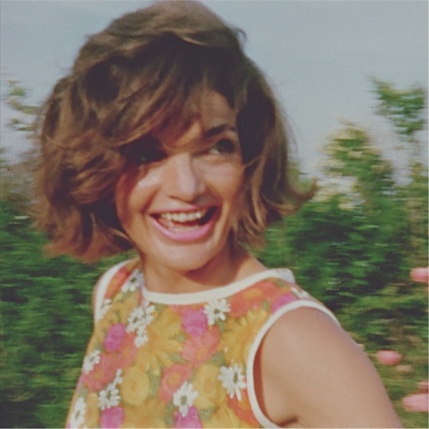 A woman smiling outdoors, wearing a colorful floral top, with short brown hair and a blurred background of greenery.